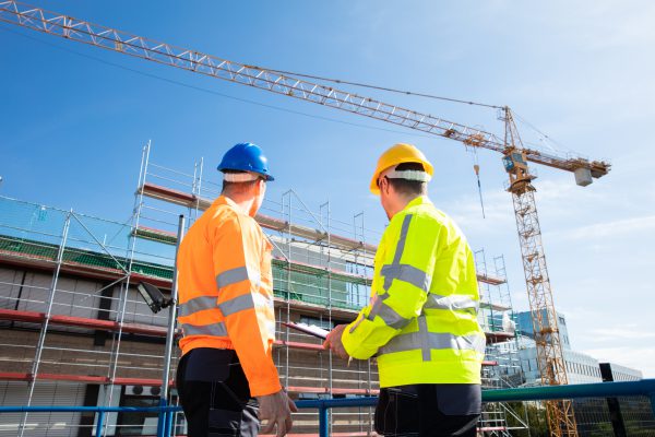 Rear,View,Of,Two,Male,Architect,Wearing,Hardhat,Looking,At