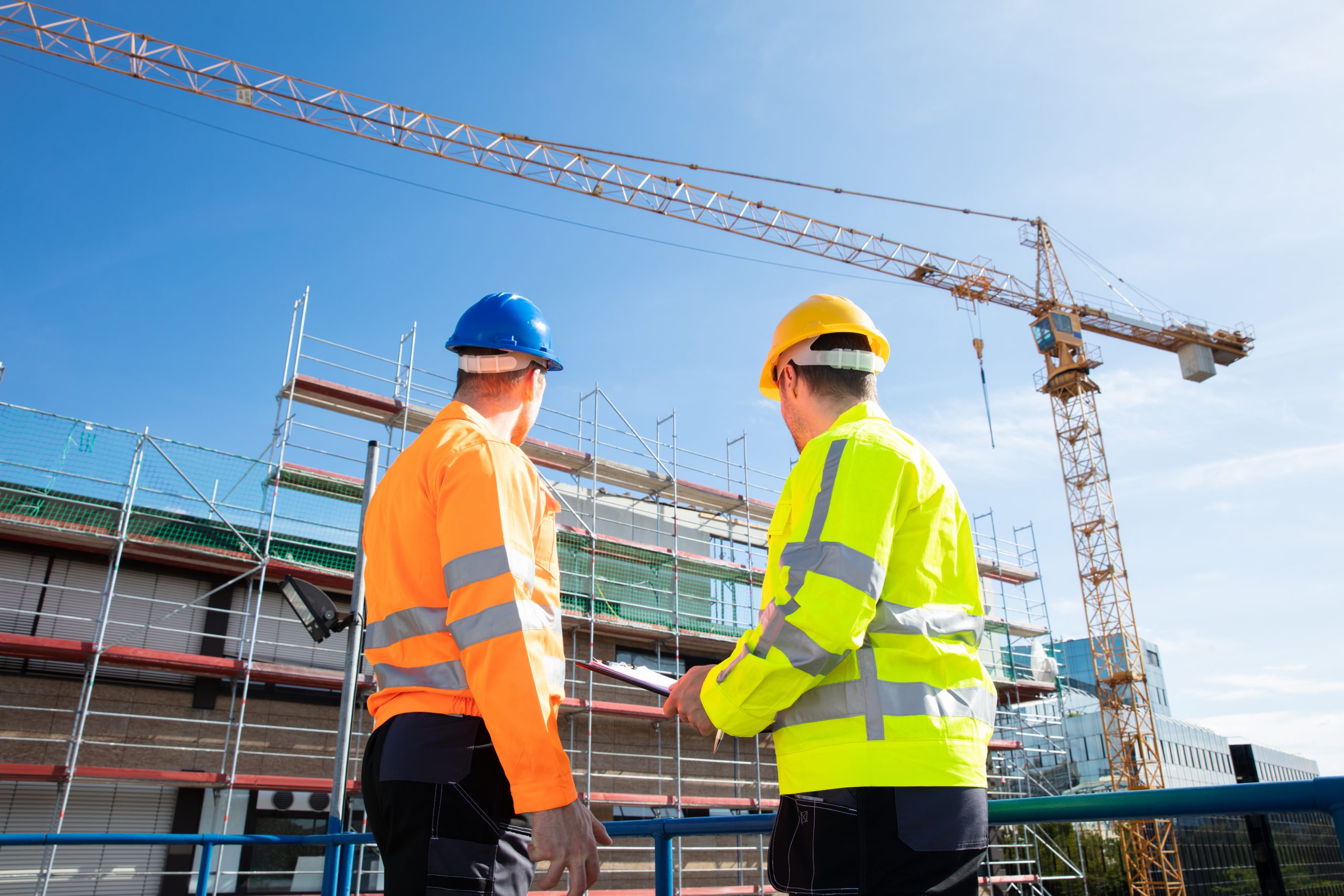 Rear,View,Of,Two,Male,Architect,Wearing,Hardhat,Looking,At